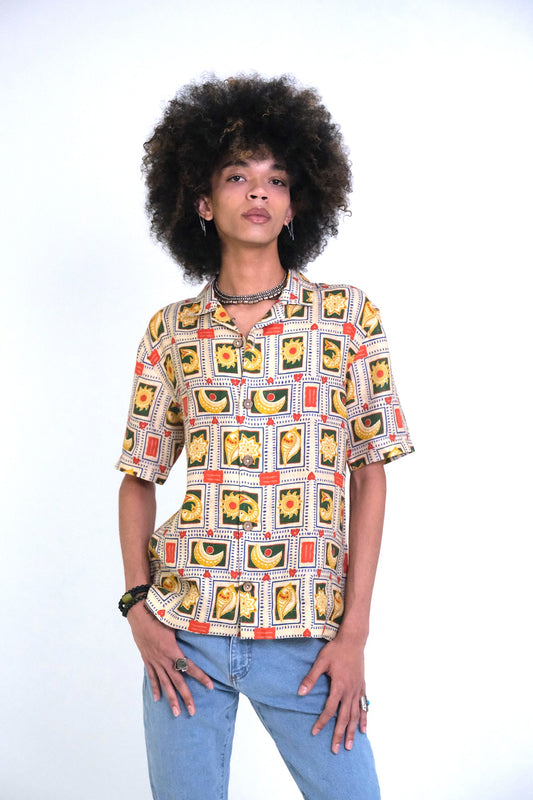 A model with an afro hairstyle wearing a RELAXED FIT short-sleeve button-up UNISEX HEMP shirt with a colorful geometric and floral print, paired with light blue jeans, against a plain white background.